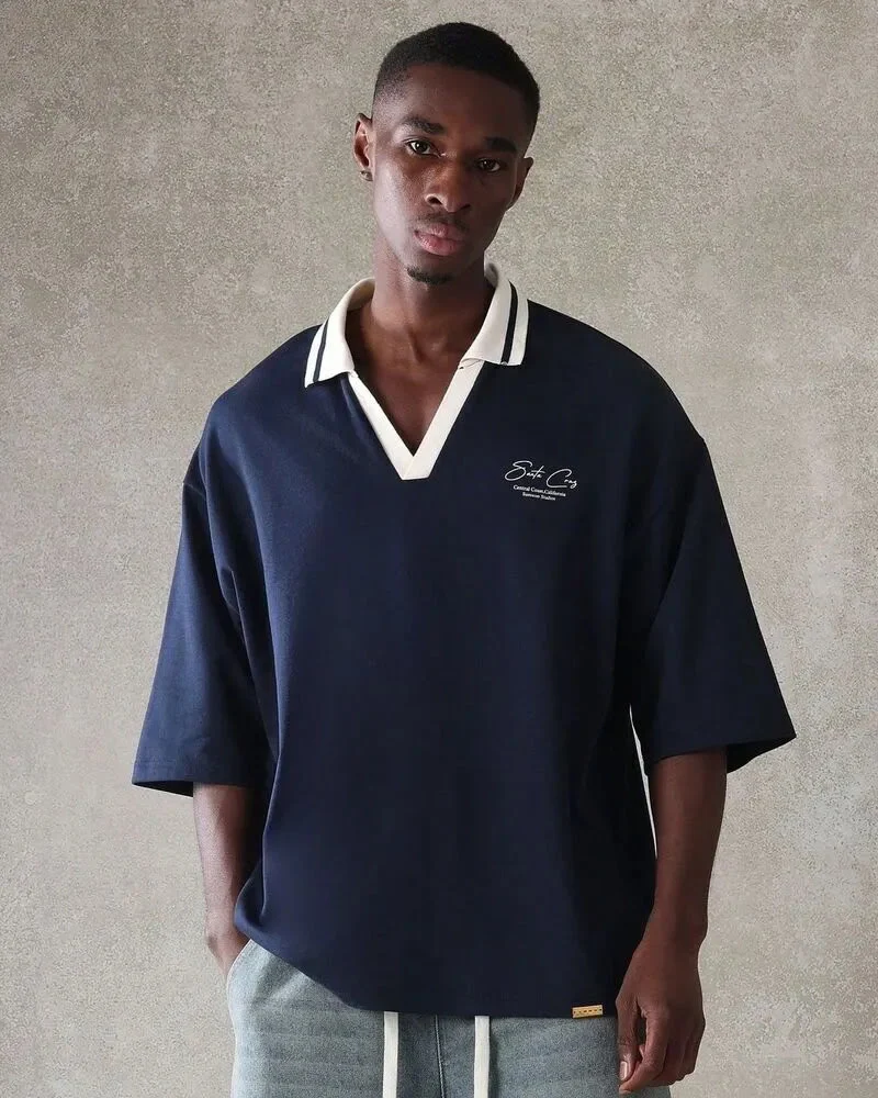 A young man posing in a navy blue oversized short-sleeved polo shirt featuring a deep V-neck white collar with navy stripes, paired with grey textured trousers.