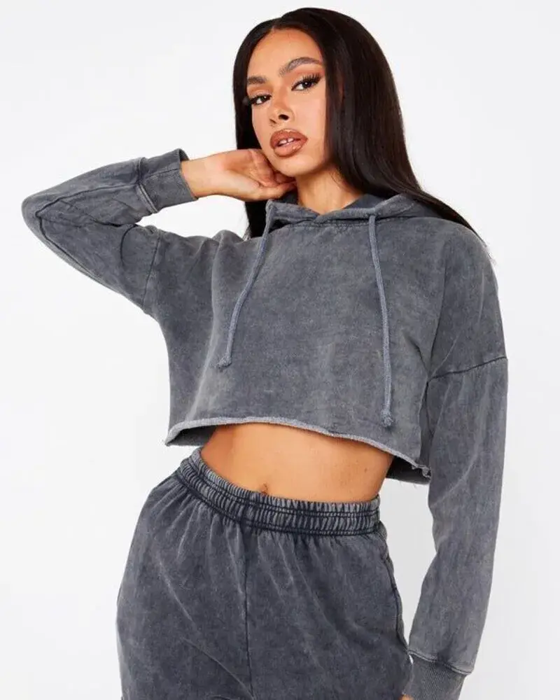 A woman posing in a dark grey acid-washed cropped hoodie with a raw hem and matching high-waisted joggers against a plain white background.
